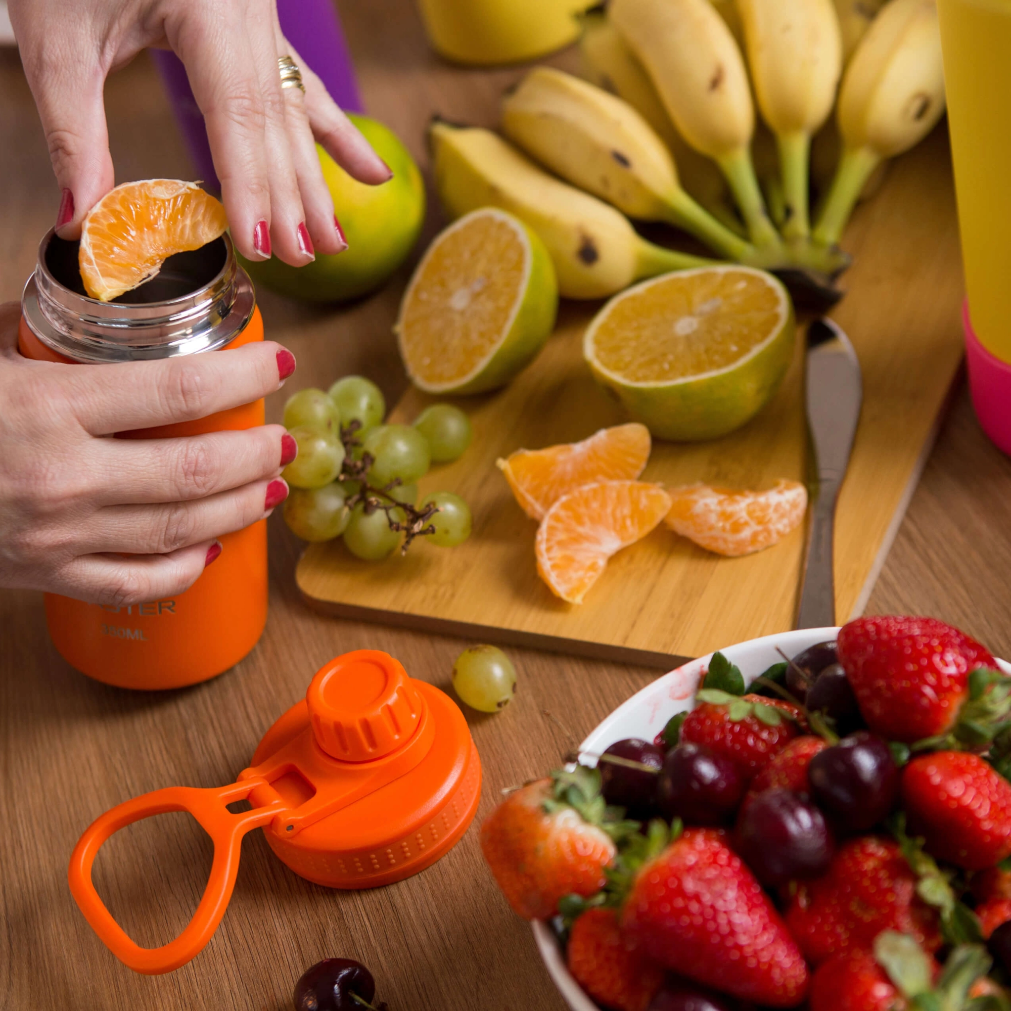 Mesa com frutas coloridas e uma mão espremendo uma laranja dentro de uma garrafa Faster